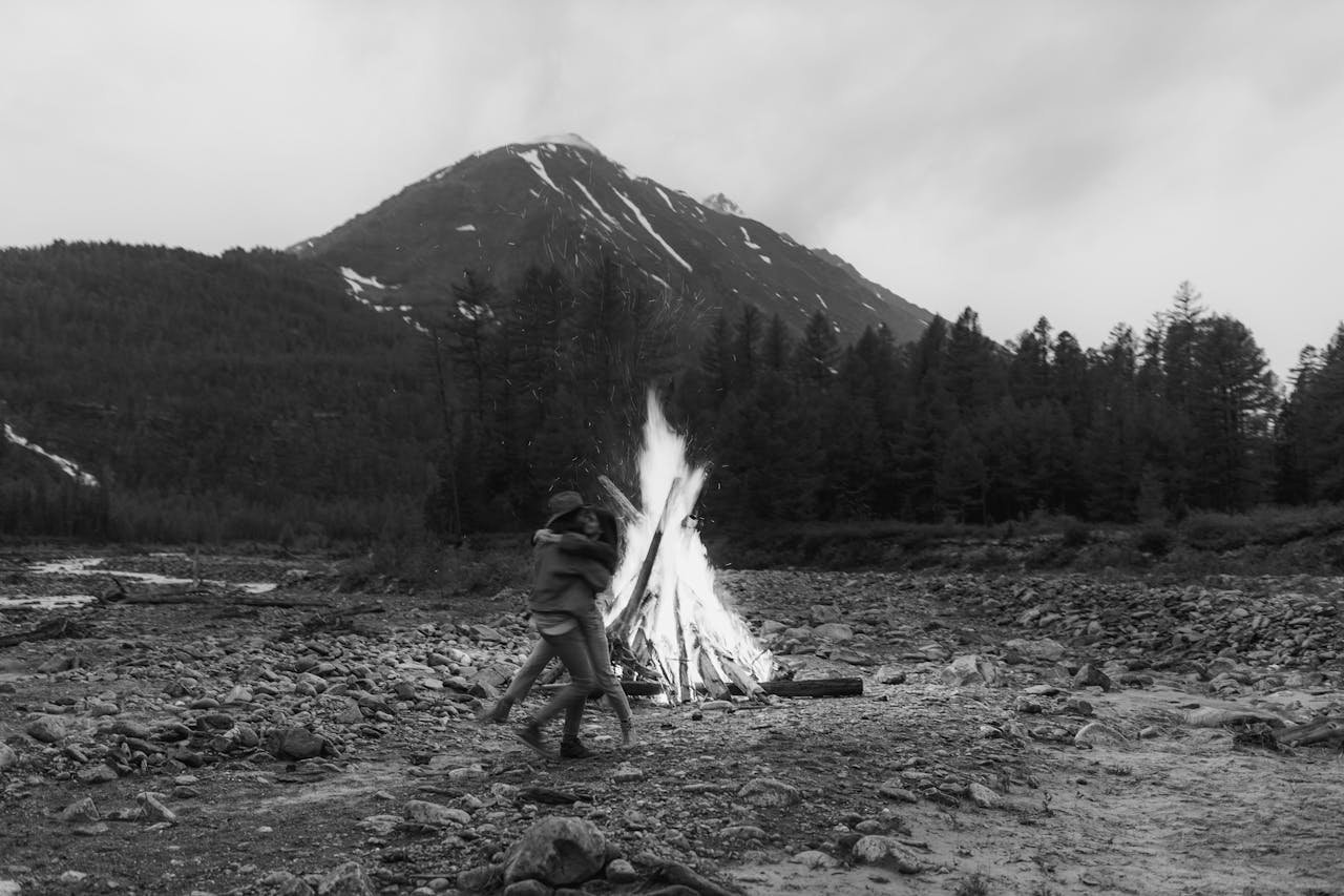 services-02 Black and white photo of a couple embracing by a bonfire with a scenic mountain backdrop.
